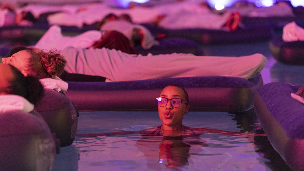 A guest floating in the warm pool under pink light at Floating Soundbath MPLS