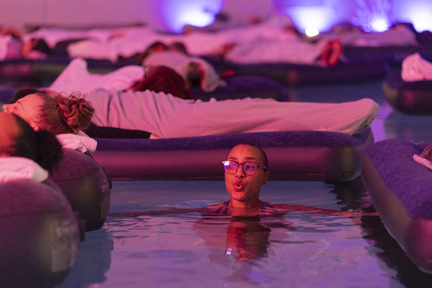 A guest floating in the warm pool under pink light at Floating Soundbath MPLS